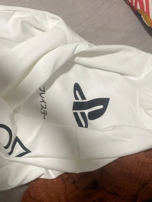 Vit PlayStation hoodie med tryck - Säljer en HELT oanvänd vit hoodie från PlayStation med det klassiska svarta PlayStation-logotypen och japansk text på bröstet. Hoodien har huva och är tillverkad i mjukt material, perfekt för gaming eller chill. Snygg och enkel design som sticker ut. Aldrig aldrig använt fick den i present men säljer nu för att den är för liten och det finns inget kvitto kvar. (STORLEK: EUR:170, US: 20, UK:14+years)