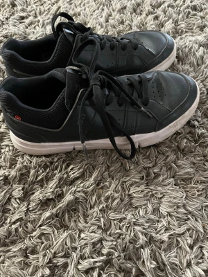 Svarta sneakers  - Stilrena svarta sneakers med vit sula och diskret logga på sidan. Skorna har klassisk låg profil, svarta skosnören och ventilerande meshdetaljer vid hälen. Ovandelen är i slätt läder och perforerade detaljer ger extra stilpoäng.