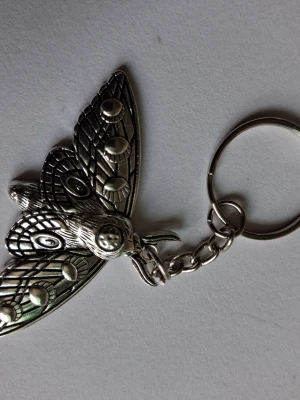 Silvrig nyckelring med nattfjäril - Unik nyckelring i metall med en detaljerad nattfjäril i silvrig färg. Fjärilen har snygga mönster på vingarna och en cool retrovibe. Perfekt att hänga på väskan eller nyckelknippan för en personlig touch.