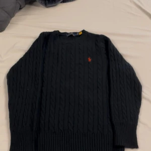 Svart kabelstickad tröja Polo Ralph Lauren - Kabelstickad/ull svart tröja från Polo Ralph Lauren med klassisk röd broderad logga på bröstet. Tröjan har rund halsringning, långa ärmar och breda ribbade muddar. Perfekt för lager-på-lager under höst och vinter.