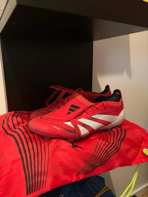 Adidas Predator Elite Fotbollskor - Snygga röda Adidas Predator fotbollsskor med vita och svarta detaljer. Skorna har klassisk snörning, låg profil och slitstark yttersula med dobbar för bra grepp på planen. Tillverkade i syntetmaterial och designade för maximal bollkontroll. Perfekta för dig som vill dominera på planen.