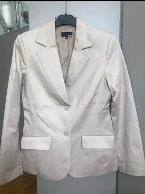 Beige kavaj från H&M - Stilren beige kavaj från H&M med klassisk krage, två knappar framtill och raka fickor. Figursydd modell med långa ärmar och slits bak. Perfekt för en clean och modern look. Materialet känns mjukt och lätt, troligen bomullsblandning.