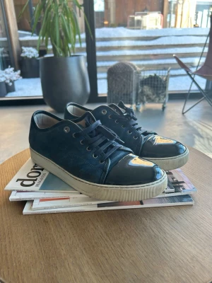 Feta Lanvin pjucks - Snygga blå sneakers från Lanvin med ovandel i mocka och lackade tåpartier. Skorna har vita sulor och matchande blå skosnören. Metallfärgade öljetter ger en clean och exklusiv look. Perfekta för dig som gillar stilrena detaljer och premiumkänsla. Skriv vid frågor!