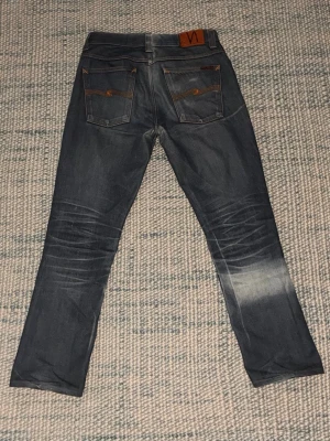 Mörkblå jeans från Nudie Jeans - Snygga mörkblå jeans från Nudie Jeans med klassisk femficksdesign och orangea sömmar. Jeansen har en rak passform och coola slitningar vid knäna och bakfickorna. Tillverkade i denim med läderpatch bak i midjan. Jeansen har professionellt sytts upp i benen från W32/L32 till ungefär W32/L30. Jeansen är också solblekta på höger ben vilket ger dem en unik look 