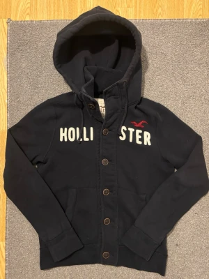 Vintage Hollister Cardigan Med Knappar - Väldigt sällsynt och vintage Hollister hoodie med knappar | Tröjan är i väldigt bra skick utan defekter! | Längd ≈ 63cm, Bredd armhåla-amrhåla ≈ 53cm | Modell 175cm, 68kg | Hör av vid ytterligare frågor och priset kan diskuteras vid snabbt köp! |