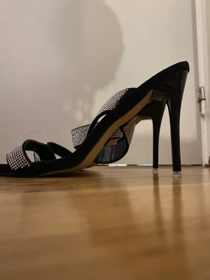 Svarta pumps med strass från Shein - Säljer ett par svarta pumps från Shein med smala klackar och två band över foten täckta av glittrande strass. Skorna har öppen tå och elegant design som ger en lyxig känsla. Perfekta för dig som vill sticka ut med lite extra bling.