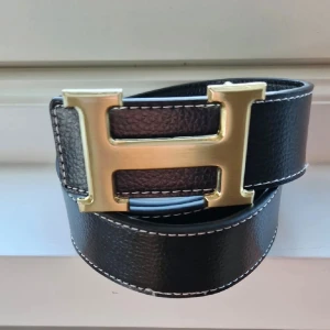 Svart skärp med guldfärgat H-spänne - Snyggt svart skärp med ett stort guldfärgat H-spänne. Skärpet är gjort i skinn med vit kontrastsöm längs kanterna och har en klassisk, stilren look som passar till det mesta.