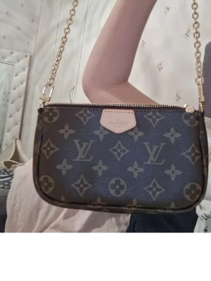  axelremsväska - Snygg handväska  brunt monogrammönster med guldiga kedjedetaljer. Väskan har en rektangulär form och dragkedja upptill samt en liten läderpatch med logga framtill. Perfekt för dig som gillar lyxiga accessoarer.