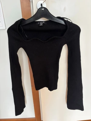 Svart stickad topp med vågig urringning - Trendig svart stickad topp från New Yorker med lång ärm och unik vågig urringning. Figurnära passform och stretchigt material som sitter snyggt på kroppen. Svårt att se på bild men den är även lite glittrig. Toppen är i Storlek S, säljer då jag inte fått användning för den. Helt oanvänd💕