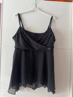Ny. Svart omlott linnetopp med volang - Snygg svart linnetopp med omlottdetalj framtill och tunna axelband. Toppen har ett luftigt, transparent tyg med volangkant nedtill och ett elegant fall. Perfekt för dig som gillar en feminin och stilren look.