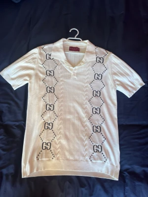 Vit kortärmad skjorta från Gucci - Snygg vit kortärmad skjorta från Gucci med klassiskt GG-mönster längs framsidan. Skjortan har krage, knappar vid halsen och är tillverkad i mjukt bomullsmaterial. Perfekt för dig som vill ha en stilren och lyxig look. Säljs då den inte används. 