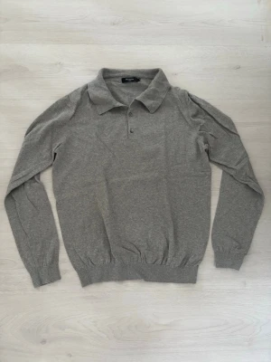 Grå långärmad tröja från Matinique - Säljer en stilren grå långärmad tröja från Matinique med klassisk krage och knappar framtill. Tröjan är tillverkad i mjukt material och har ribbade muddar vid ärmslut och nederkant. Perfekt för en enkel outfit med en vit tröja under och ett par jeans.