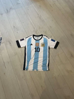 Argentina Messi matchtröja blåvit - Säljer en officiell Argentina matchtröja från Adidas med Messi och nummer 10. Tröjan har klassiska blå och vita ränder, svarta detaljer och broderade märken på bröstet. Korta ärmar och rund hals. Perfekt för fotbollsfans!