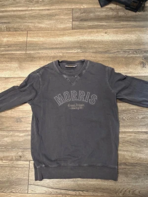 Grå tröja från Morris - Snygg grå sweatshirt från Morris med tryckt logga och text på bröstet. Tröjan har rund halsringning, ribbade muddar vid ärmslut och nederkant samt avslappnad passform. Perfekt för en casual look.