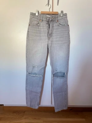 Gråa högmidjade ”mom” jeans med slitningar - Säljer ett par ljusgrå ”mom” jeans med hög midja och raka ben. Jeansen har slitna detaljer och hål vid knäna för en trendig, avslappnad look. Klassisk femficksmodell och stängs med knapp och dragkedja. Perfekta för en cool och avslappnad stil.