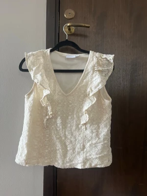 Vit spetsblus med volangdetaljer - Söt vit blus i spets med v-ringning och volangdetaljer längs axlarna. Ärmlös modell som ger en luftig känsla och passar perfekt till sommaren. Blusen har ett romantiskt mönster och är fodrad undertill för extra komfort. Det har gått en tråd men går lätt att sy ihop. Säljer för att den är för liten. Från Vila. Storlek 34.
