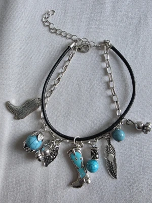 Armband med turkosa pärlor och berlocker - Coolt armband med silverfärgade kedjor och svart läderband. Utsmyckat med turkosa pärlor och berlocker i form av stövel, fjäder, vinge och andra detaljer. Perfekt accessoar för en unik stil.