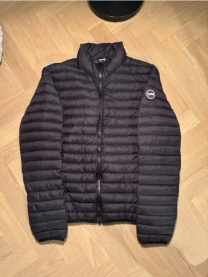 Svart dunjacka från Colmar - Snygg svart dunjacka från Colmar med quiltad design och hög krage. Jackan har dragkedja framtill och en rund Colmar-logga på vänster ärm. Perfekt för kalla vinterdagar och ger en stilren look.