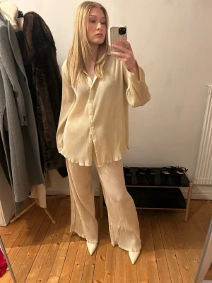 Ljusgult plisserat set med skjorta och byxa - Säljer ett gult plisserat set med lös passform. Setet består av en långärmad skjorta med krage och knappar framtill samt matchande vida byxor med resår i midjan. Materialet är lätt och luftigt, perfekt för en avslappnad men stilren look. Passar för personer i längden 1,65-175. Högmodjade byxor men går att vika till lågmidjade om önskat. För en ”lose fit” Small eller M för en tajtare L.