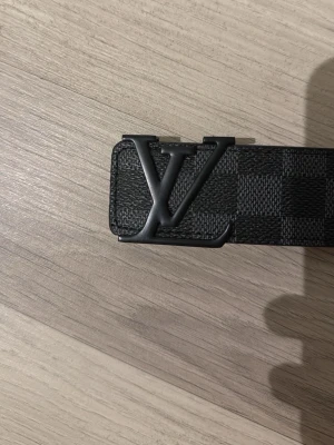 Svart rutigt LV-bälte - Snyggt svart bälte från Louis Vuitton med klassiskt rutigt mönster och stor LV-logga i metall på spännet. Bältet har en stilren design och passar perfekt till jeans eller kostymbyxor. Materialet är skinn med en exklusiv känsla.