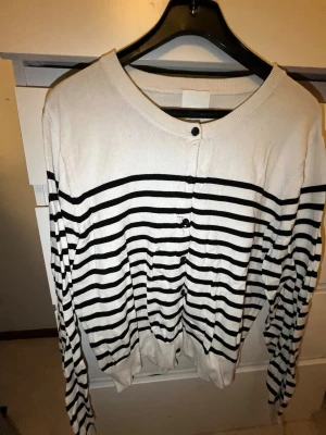 Randig vit och svart kofta - Kofta i vitt med svarta horisontella ränder och rund halsringning. Långa ärmar och knäppning framtill med svarta knappar. Tunn och mjuk kvalitet, perfekt för lager på lager. Klassisk och enkel design som passar till det mesta.