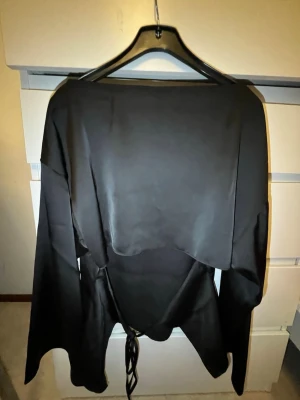Svart omlottopp med snörning - Snygg svart omlottopp i satinliknande material med långa ärmar och snörning i midjan. Toppen har en elegant, lös passform och en lätt glansig finish som ger en lyxig känsla. Perfekt för dig som vill ha något stilrent och trendigt.