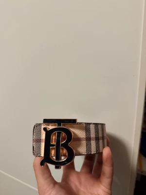 Rutig beige Burberry skärp - Snyggt rutig beige och brun Burberry skärp med ikoniskt B-spänne i guld och svart. Bältet har klassiskt rutmönster och bred design som gör det till en statement-accessoar. Perfekt för att lyfta vilken outfit som helst.