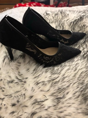 Svarta spetsiga pumps med spetsdetaljer - Säljer ett par svarta klackar  med spetsig tå och snygga spetsdetaljer på sidorna. Klackarna är smala och eleganta, vilket ger en stilren look. Materialet är en mix av tyg och spets som ger skorna en unik och trendig känsla. Dom har bara används 2 gånger men säljer dom för dom är för stora 