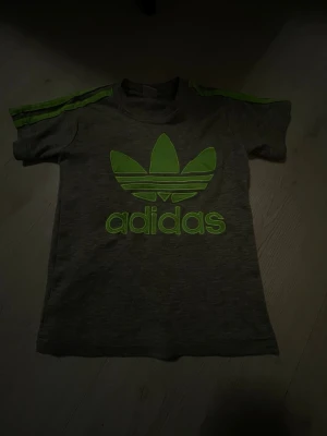 Grå Adidas t-shirt med grönt tryck - Snygg grå t-shirt från Adidas med stort gröna  ränder på axlarna. Klassisk passform och rund hals.