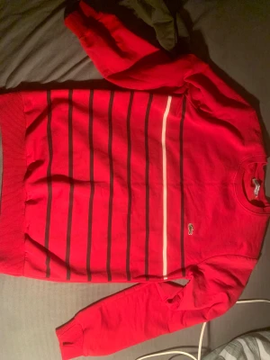 Röd randig tröja från Lacoste M passar också s - Säljer en röd tröja från Lacoste i storlek M med svarta och en vit rand över bröstet. Klassisk krokodil-logga broderad på vänster sida. Tröjan har rund hals, ribbade muddar och är långärmad. Perfekt för dig som gillar sportig och stilren look. Ge bud