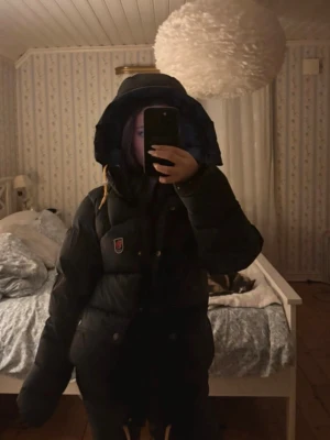 Svart dunjacka med huva från Fjällräven - Säljer en svart dunjacka från Fjällräven med stor, varm huva och klassisk logotyp på bröstet. Jackan har knappar och dragkedja framtill samt två stora fickor. Perfekt för kalla vinterdagar och riktigt mysig att bära.
