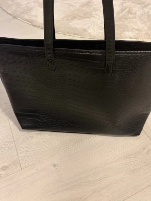 Svart shoppingväska i krokopräglat material - Stilren svart shoppingväska med krokodilpräglat mönster och dubbla axelband. Väskan har en fyrkantig form och är rymlig nog för allt du behöver. Perfekt för dig som gillar en trendig och klassisk look. Materialet är konstläder.