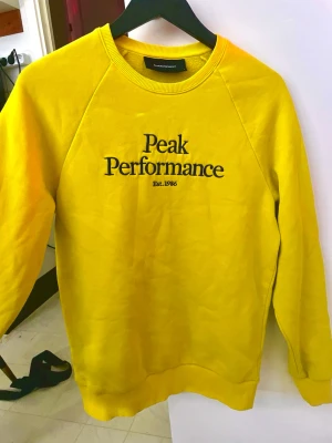 Peak Performance Gul Sweatshirt - Gul sweatshirt från Peak Performance med svart broderad logga på bröstet. Klassisk rund halsringning och ribbade muddar vid ärmslut och nederkant. Perfekt för en avslappnad och sportig stil.
