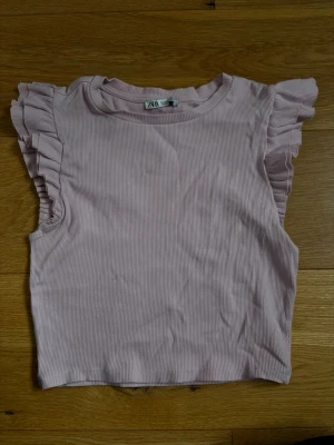 Ljusrosa crop top med volangärmar - Supersöt ljusrosa crop top från Zara med ribbad struktur och volangdetaljer på ärmarna. Toppen har rund halsringning och är ärmlös med breda volanger som ger en trendig och feminin look. 