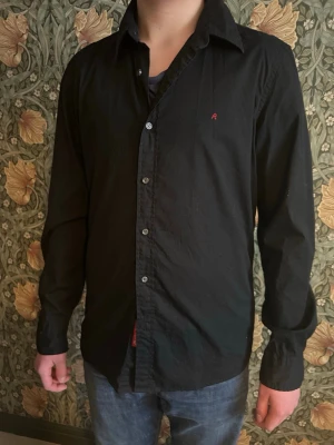 Svart skjorta från Replay Blue Jeans - Snygg svart skjorta från Replay Blue Jeans med klassisk krage och knappar framtill. Liten röd logga på bröstet ger en clean detalj. Skjortan är långärmad och gjord i mjuk bomull, perfekt till jeans eller chinos. Slim large 