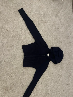 Mörkblå hoodie från Gina Tricot - Mörkblå hoodie från Gina Tricot med dragkedja framtill och huva. Tröjan har långa ärmar och ribbade muddar vid midjan. Perfekt för en avslappnad och cool stil.