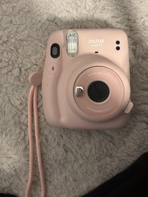 Rosa Instax Mini 11 kamera - Säljer en rosa Instax Mini 11 direktfilmskamera med rundad form och matchande handledsrem. Kameran har inbyggd blixt, enkel knapp för att ta bilder och klassisk Instax-design. Perfekt för att få ut fysiska bilder direkt. Tillverkad i plast.