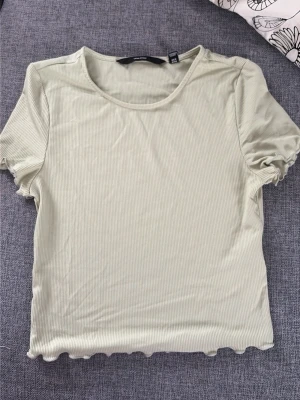 Ljusgrön ribbad topp från Vero Moda - Snygg ljusgrön ribbad topp från Vero Moda i storlek XS. Kortärmad modell med croppad passform och vågig kant nertill. Tillverkad i mjuk och stretchig bomullsmix, perfekt till jeans eller kjol.