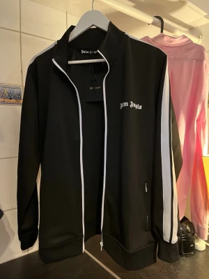 Svart track jacket från Palm Angels - Snygg svart track jacket från Palm Angels med vita ränder längs ärmarna och vit dragkedja. Jackan har ståkrage, fickor med dragkedja och logga broderad på bröstet. Perfekt för en sportig och trendig streetwear-look. Storlek Medium  bara att höra av sig ifall man har nån fråga