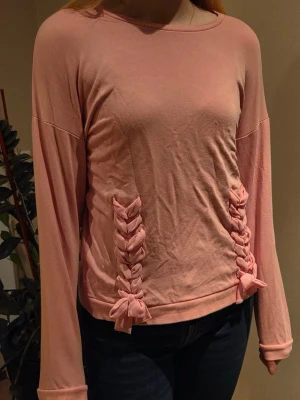 Rosa tröja med flätade detaljer Lindex - Söt ljusrosa långärmad tröja från Lindex med unika flätade banddetaljer framtill. Tröjan har rund halsringning och är gjord i mjukt, stretchigt material som känns skönt mot huden. Perfekt för dig som gillar en gullig och trendig stil.