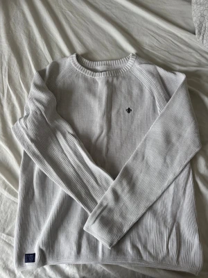 Vit stickad tröja från Morris (M) - Snygg vit sällsynt stickad tröja från Morris med diskret logga broderad på bröstet. Tröjan har rund halsringning, ribbade muddar och är tillverkad i ett mjukt, strukturerat material. Perfekt för dig som gillar stilrena och klassiska plagg.