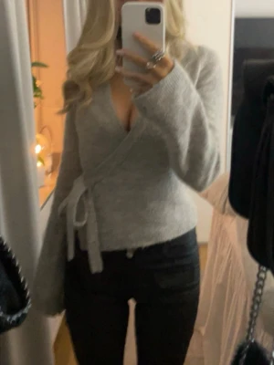 Grå omlottkofta med knytband - Mysig grå omlottkofta med knytband i sidan och långa ärmar. Koftan har en djup v-ringning och är tillverkad i ett mjukt stickat material som passar perfekt för lager-på-lager. Enkel att matcha med jeans eller kjol.