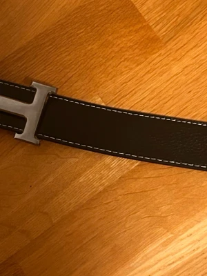 Svart skärp mer h3rm3s  - Snyggt svart skärp i skinn med vit kontrastsöm och ett stort h märke metallspänne. Perfekt accessoar för att lyfta din outfit med en stilren och klassisk touch.