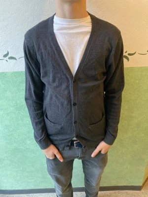 Mörkgrå Riley  kofta med knappar - En stilren mörkgrå kofta med v-ringning och knäppning framtill. Koftan har två fickor och långa ärmar. Perfekt att slänga över en t-shirt för en enkel och clean look.