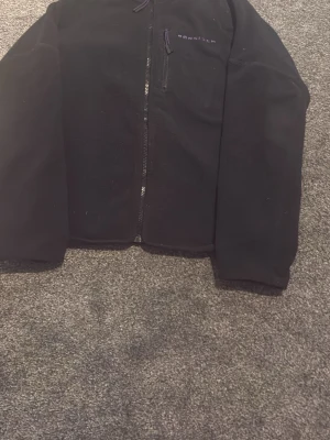 Svart fleecejacka från Röhnisch - Mysig svart fleecejacka från Röhnisch med dragkedja framtill och en bröstficka med lila detalj. Jackan har lång ärm och enkel, stilren design med broderad logga på bröstet. Perfekt för lager på lager under kyliga dagar.