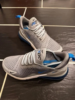 Nike Air Max 270 grå/blå sneakers - Nike Air Max 270 sneakers i grå mesh med blå detaljer och synlig Air-enhet i hälen. Skorna har en sportig siluett, ventilerande ovandel och klassisk Nike-logga på sidan och plösen. Perfekta för dig som gillar streetwear och bekväma sneakers.