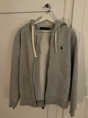Grå zip hoodie från Polo Ralph Lauren - Snygg grå hoodie med dragkedja från Polo Ralph Lauren. Klassisk design med huva och vita snören, två fickor framtill och den ikoniska lilla loggan broderad på bröstet. Tillverkad i mjuk bomullsblandning som är skön att ha på sig.