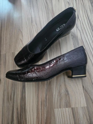 Vinröda pumps med krokopräglat mönster - Säljer ett par vinröda pumps från ara med glansig krokopräglad yta och låg klack med gulddetalj. Skorna har en klassisk, fyrkantig tå och är tillverkade i syntetmaterial som ger en lyxig känsla. Perfekta för dig som gillar tidlös och elegant stil. Innermått 22.5 . Strl 37.