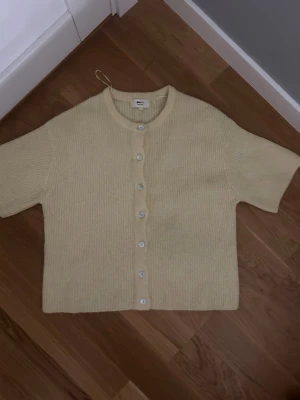 Ljusgul stickad kofta från Gina Tricot - Kortärmad stickad kofta i ljusgul ullblandning från Gina Tricot. Koftan har rund halsringning, ribbstickad struktur och vita knappar framtill. Perfekt för lager-på-lager och en stilren look.