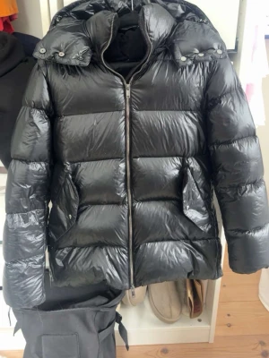 Svart pufferjacka med huva Massimo dutti  - Säljer en svart pufferjacka med glansig finish och stor huva. Jackan har dragkedja framtill, två stora fickor med lock och quiltad design. Perfekt för kalla vinterdagar och riktigt skön att bära. Passar dig som gillar en stilren och enkel look.